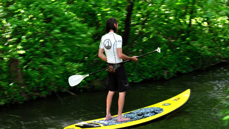 Stand up paddling in the climbing park, © Soulriders Person stand up paddling on a river, surrounded by green vegetation.