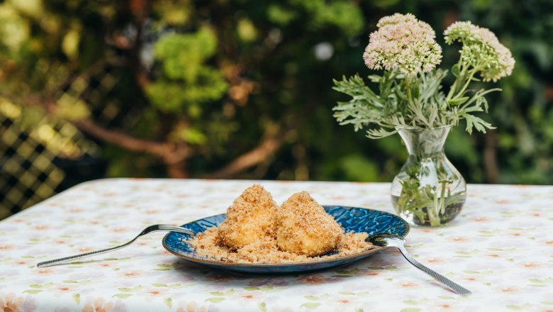 Zwetschkenknödel, © Niederösterreich Werbung/Daniela Führer Ein Teller mit zwei Zwetschkenknödeln auf einem Tisch mit Blumenmusterdecke, daneben eine Vase mit Blumen.