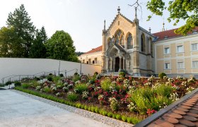 KirchemitGartenanlage, © SusanneHammerle Eine Kirche mit gotischen Elementen und einem gepflegten Garten voller Blumen im Vordergrund.