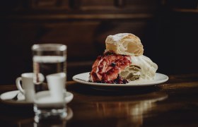 Klostercremeschnitte, © Niederösterreich Werbung/Sophie Menegaldo Ein Teller mit einer Klostercremeschnitte, garniert mit roten Beeren, steht auf einem Holztisch. Im Hintergrund sind ein Glas Wasser und zwei Espressotassen zu sehen.