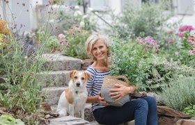 Sommergarten Christines Paradies, © Eva Lekey Fotografie Eine Frau sitzt mit einem Hund in einem blühenden Garten auf Steinstufen.