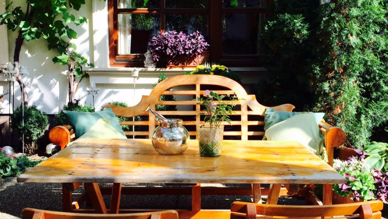 Heurigen Schwertführer, © Schwertführer Gemütlicher Gartenbereich mit Holztisch und Stühlen, umgeben von Pflanzen und Weinreben.