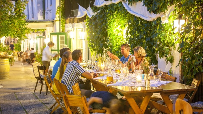 Gebeshuber, © Gebeshuber Menschen sitzen an einem langen Tisch im Freien, umgeben von Pflanzen und Lichtern.