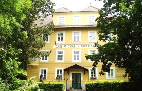 Frontansicht, © Carl-Michael-Zieherer Haus Frontansicht des Carl Michael Ziehrer-Hauses, ein gelbes Gebäude mit mehreren Fenstern und einem grünen Eingang, umgeben von Bäumen.