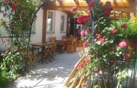 Rosenbogen, © Dominik Herzog Ein Rosenbogen mit roten Rosen führt zu einem Außenbereich mit Holztischen und -stühlen.