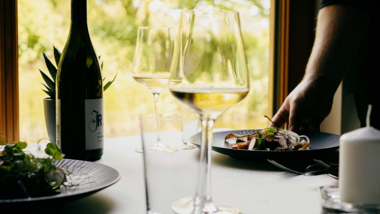 Restaurant im Weingut Johnneshof Reinisch, © Julius Hirtzberger Ein gedeckter Tisch mit Weinflasche, Weingläsern und einem Teller mit Essen in einem Restaurant.