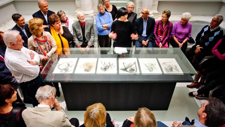 Exhibition tour, © Arnulf Rainer Museum, Foto: Kollektiv Fischka A group of people stand around a glass table with drawings in an exhibition room.