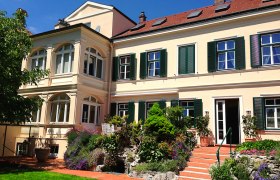 Hausansicht, © Helmut Förster Ein großes, zweistöckiges Haus mit gelber Fassade und grünen Fensterläden, umgeben von einem gepflegten Garten mit Blumen und Bäumen.