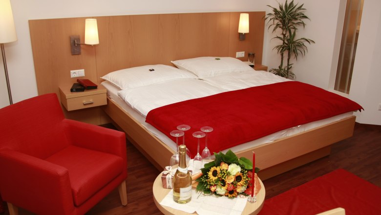 Doppelzimmer, © City Hotel Mödling Ein gemütliches Doppelzimmer mit rotem Sessel, Blumenstrauß und Sekt auf einem Tisch.