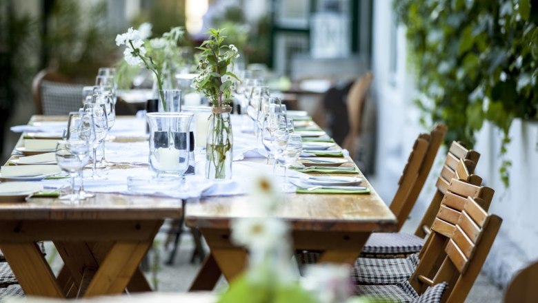 Gebeshuber, © Gebeshuber Langer Holztisch im Freien, gedeckt mit Gläsern, Besteck und Blumen.