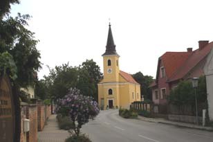 Günselsdorf, © Weinpanorama Eine gelbe Kirche mit Turm in einer ruhigen Straße, umgeben von Bäumen und Häusern.