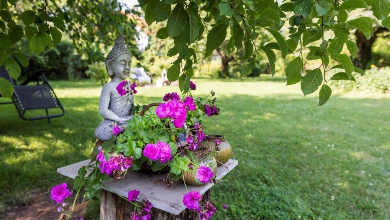 Rosenbauchs Restaurantgarten, © Karl Rosenbauch Garten mit Buddha-Statue und pinken Blumen auf einem Holzstumpf.