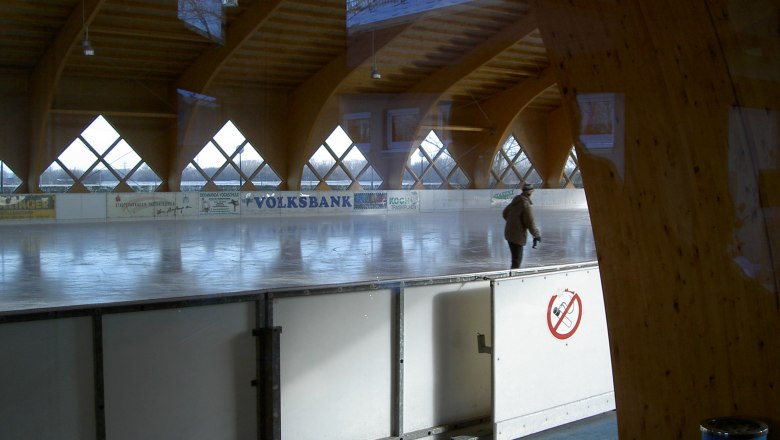Überdachte Kunsteisbahn, © Kunsteisbahn Traiskirchen Eine überdachte Kunsteisbahn mit einer Person, die darauf Schlittschuh läuft. Die Bahn ist von Holzbalken und Fenstern umgeben.