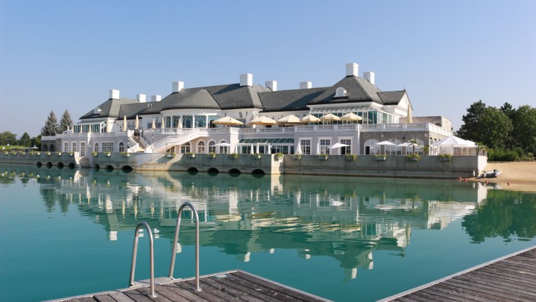 An elegant clubhouse on the lake with a terrace and sunshades, surrounded by clear water and a jetty in the foreground.