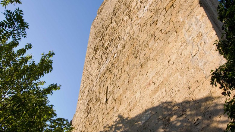 Close-up of a tall, old stone castle tower, surrounded by trees, under a blue sky.