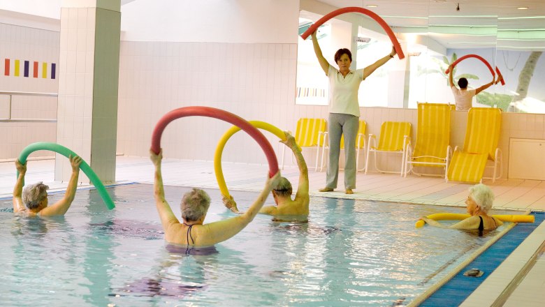 Seniorengruppe bei Wassergymnastik im Hallenbad mit Schwimmnudeln.