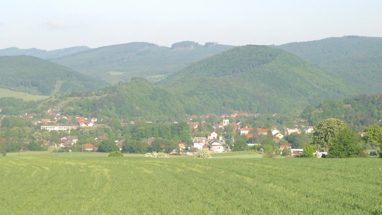 Landschaftsansicht von Alland mit gr&uuml;nen Feldern und bewaldeten H&uuml;geln im Hintergrund.