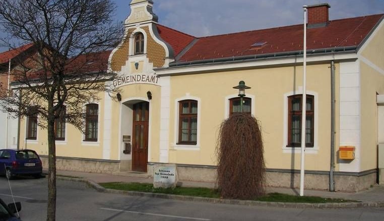 Gelbes Gebäude mit der Aufschrift 'Gemeindeamt', rotes Dach, Baum und Laterne davor.
