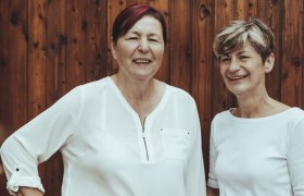 Zwei Frauen in wei&szlig;en Blusen stehen vor einer Holzwand und l&auml;cheln.