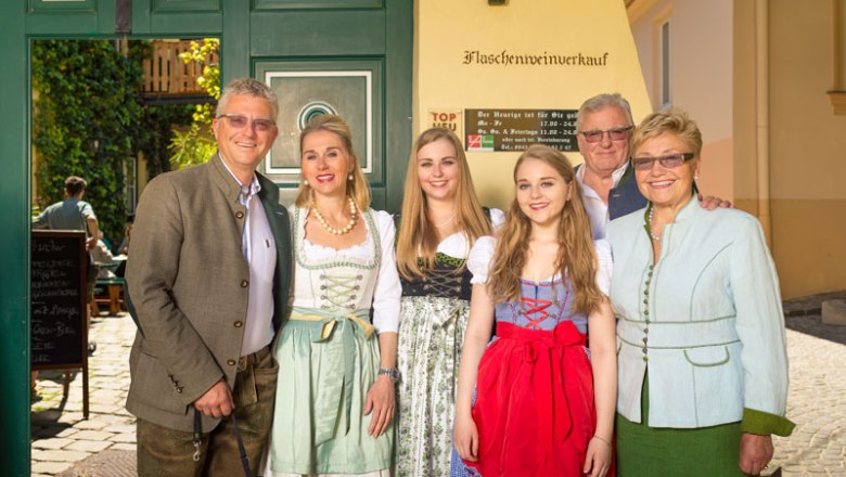 Eine Familie in traditioneller Tracht vor einem Gebäude mit der Aufschrift 'Flaschenweinverkauf'.