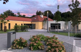 Modernes Geb&auml;ude mit gelber Fassade und rotem Dach, umgeben von Pflanzen und B&auml;umen, bei Sonnenuntergang.