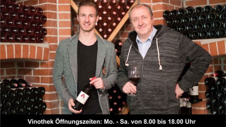Zwei M&auml;nner in einer Weinkellerei, umgeben von Weinflaschen, einer h&auml;lt ein Glas Wein.