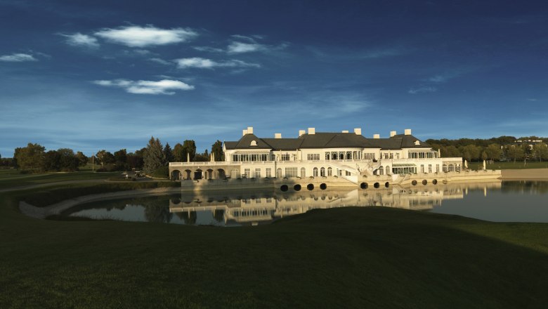 Ein elegantes Clubhaus spiegelt sich in einem ruhigen See, umgeben von grünen Rasenflächen und Bäumen unter einem klaren blauen Himmel.
