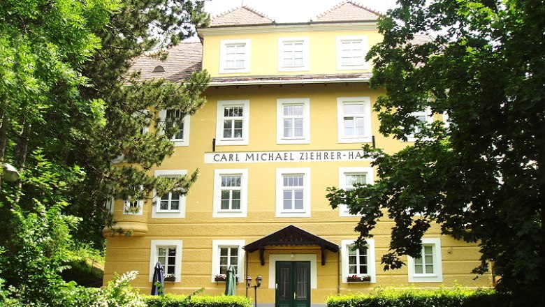 Front view, © Carl-Michael-Zieherer Haus Front view of the Carl Michael Ziehrer House, a yellow building with several windows and a green entrance, surrounded by trees.