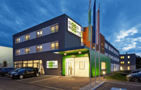 Modernes Gebäude mit der Aufschrift 'Good Rooms' bei Dämmerung, umgeben von Autos und Fahnen.