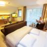 Modern hotel room with double bed, desk, TV and a woman reading the newspaper.