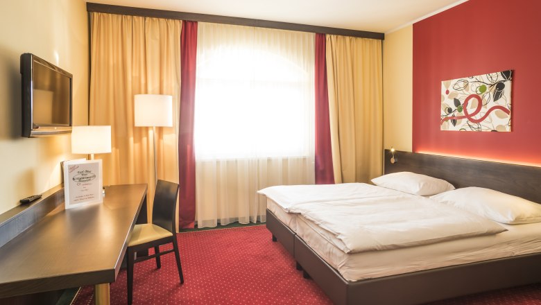 Ein gemütliches Hotelzimmer mit Doppelbett, rotem Teppich, Schreibtisch, Fernseher und moderner Kunst an der Wand.