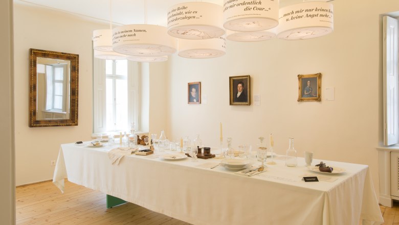 Ein gedeckter Tisch in einem Raum mit Porträts an der Wand und hängenden Lampen mit Texten.