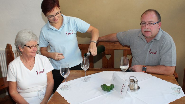 Drei Personen sitzen an einem Tisch, eine Frau schenkt Wein ein.