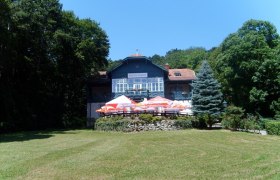 Ein gro&szlig;es Haus namens Rudolfshof mit Terrasse und Sonnenschirmen, umgeben von B&auml;umen und einer Wiese.