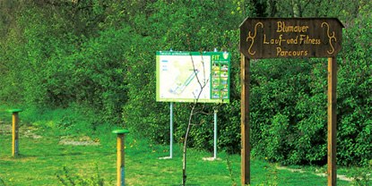 Holzschild mit der Aufschrift 'Blumauer Lauf- und Fitness Parcours' vor einer Infotafel im Grünen.