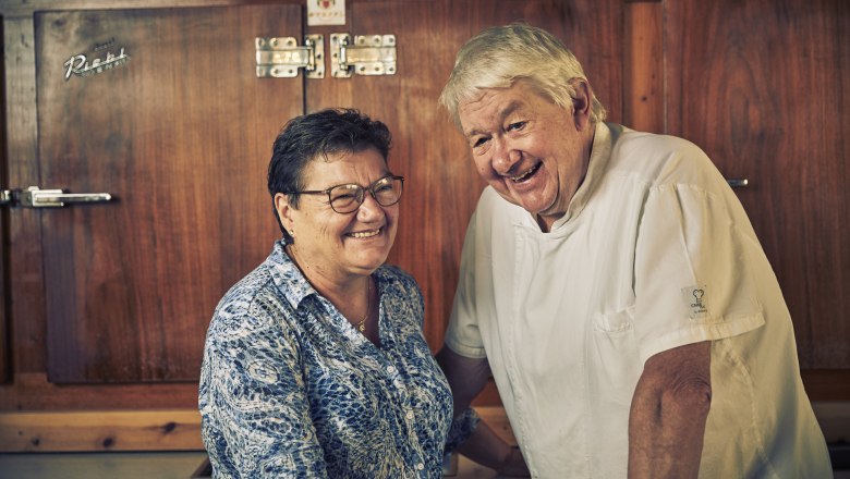 Ein älteres Paar lächelt in einer Küche. Der Mann trägt eine Kochjacke, die Frau eine gemusterte Bluse. Sie stehen vor einem Holzschrank.