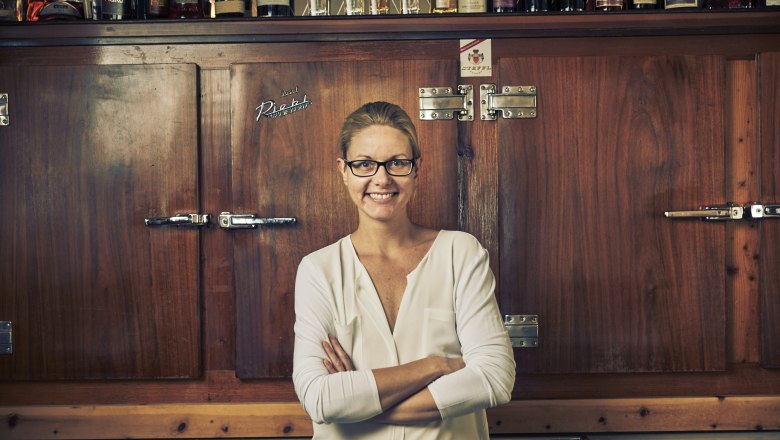 Eine Frau mit Brille steht lächelnd vor einem Holzschrank mit Flaschen darauf.