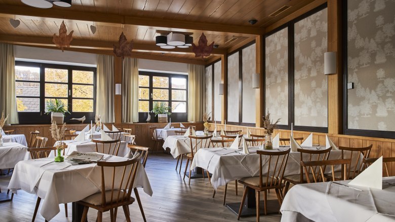 Ein elegantes Restaurant mit Holzmöbeln, weißen Tischdecken und großen Fenstern, die herbstliche Bäume zeigen.