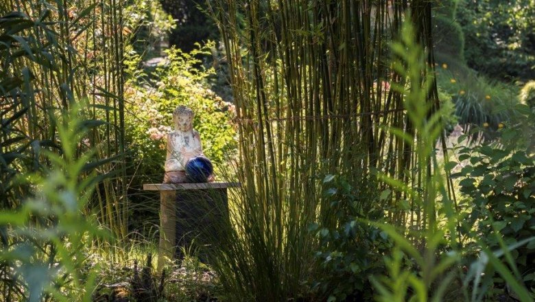 Ein asiatisch inspirierter Garten mit einer Buddha-Statue auf einem Tisch, umgeben von Bambus und &uuml;ppigem Gr&uuml;n.