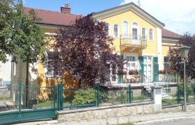 Gelbes Haus mit Balkon und grünem Zaun, umgeben von Bäumen.
