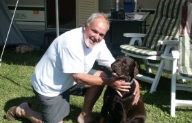 Ein Mann kniet neben einem braunen Hund auf einer Wiese vor einem Wohnwagen.