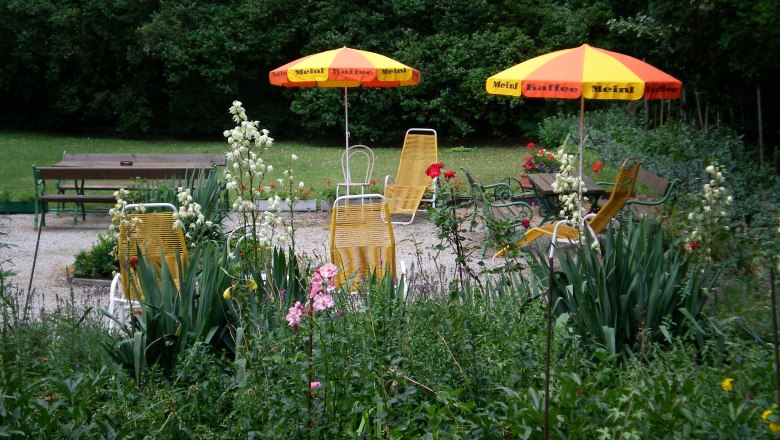 Garten mit gelben Liegestühlen und Sonnenschirmen, umgeben von Blumen und Bäumen.