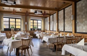 Ein elegantes Restaurant mit Holzmöbeln, weißen Tischdecken und großen Fenstern, die herbstliche Bäume zeigen.