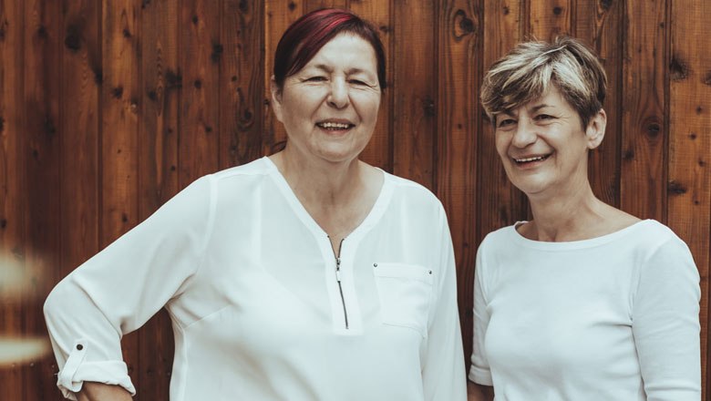 Zwei Frauen in wei&szlig;en Blusen stehen vor einer Holzwand und l&auml;cheln.