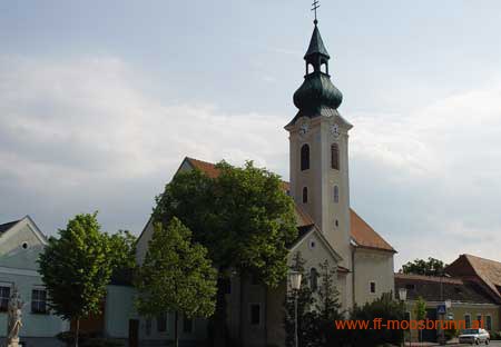 Kirche in Moosbrunn mit gr&uuml;nem Kirchturm und umliegenden Geb&auml;uden.
