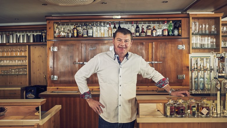 Ein Mann in einem weißen Hemd steht hinter einer Bar mit Flaschen und Gläsern im Hintergrund.