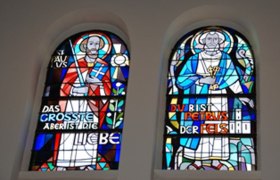 Zwei bunte Kirchenfenster mit religiösen Figuren und Inschriften.