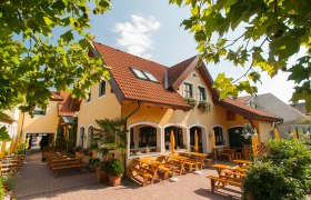 Ein gelbes Geb&auml;ude mit roten Dachziegeln und einem Biergarten mit Holzb&auml;nken und Sonnenschirmen.