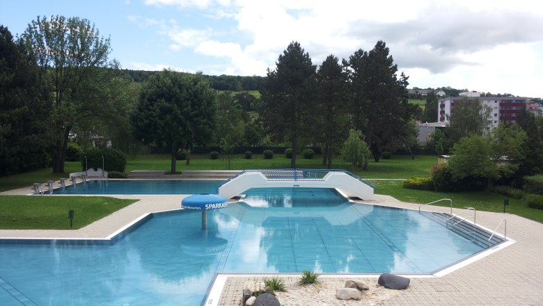 Ein leeres Freibad mit mehreren Becken, umgeben von Bäumen und Rasenflächen, unter einem bewölkten Himmel.
