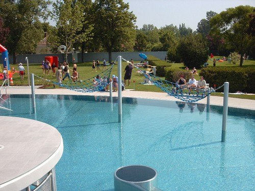 Ein Freibad mit H&auml;ngematten &uuml;ber dem Wasser und Menschen im Hintergrund auf der Liegewiese.